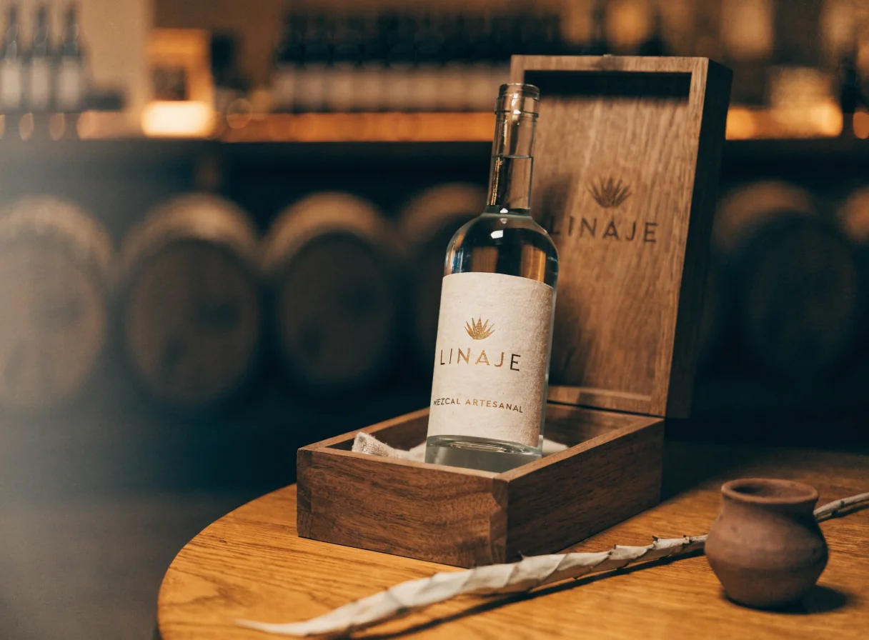 caja de nogal grabada abierta con una botella de mezcal artesanal de la marca Linaje parada dentro, etiqueta con emblema de agave, luz cálida de mesa de cata y barricas desenfocadas al fondo
