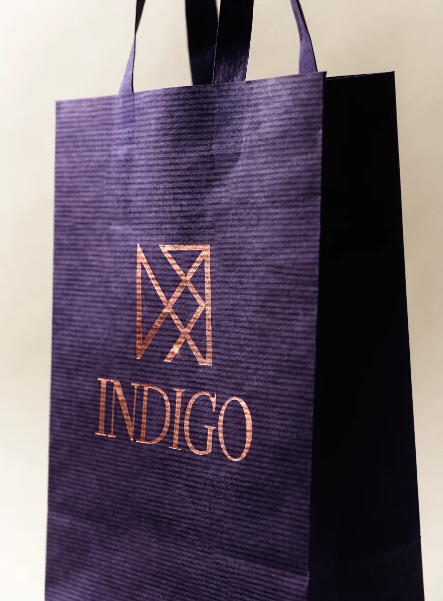 bolsa de papel kraft color índigo con fuelle lateral y la palabra INDIGO estampada en foil cobre bajo un monograma, con asas de cinta, sobre fondo crema con luz cálida