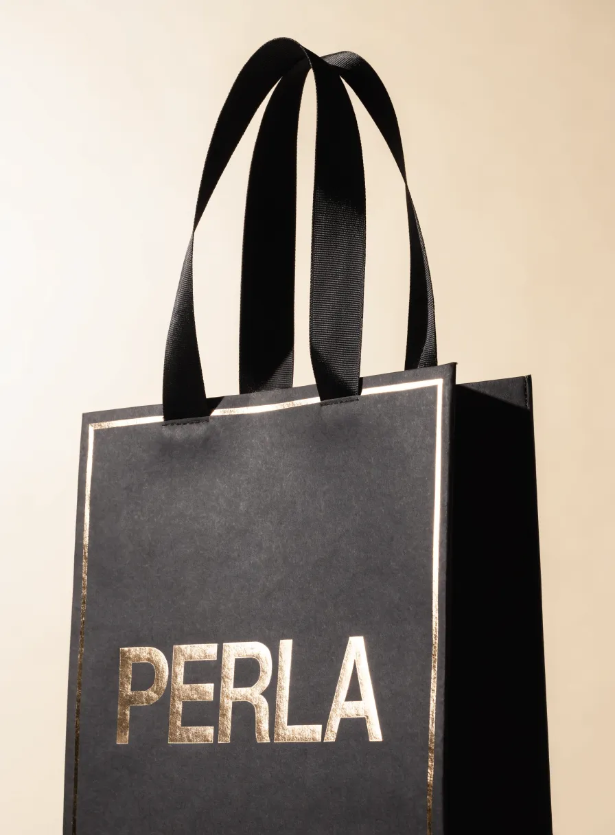 bolsa de papel laminado negro mate con la palabra PERLA y un filo en foil oro, con asas de listón grosgrain negro, sobre fondo crema con luz cálida