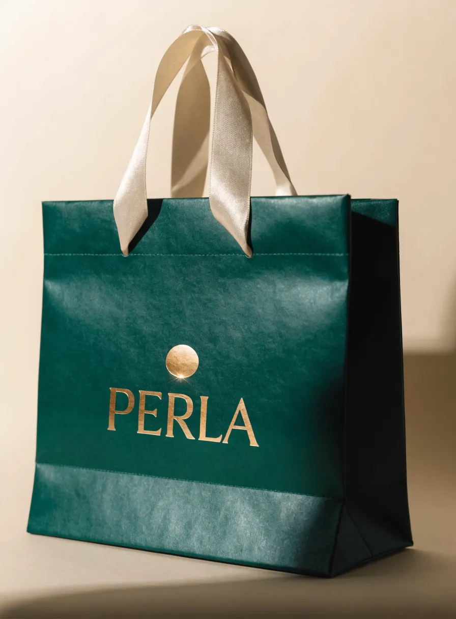 bolsa de papel laminado verde esmeralda con la palabra PERLA en foil oro bajo un emblema de perla, con asas de listón de satín marfil, sobre fondo crema con luz cálida