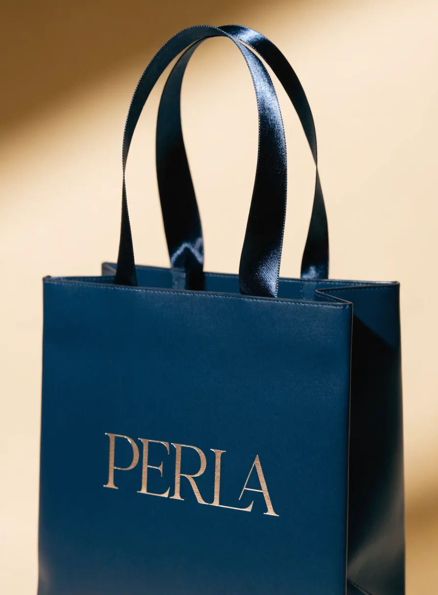 bolsa de papel laminado en azul marino con la palabra PERLA en foil champán y asas de listón de satín, sobre fondo crema con luz cálida