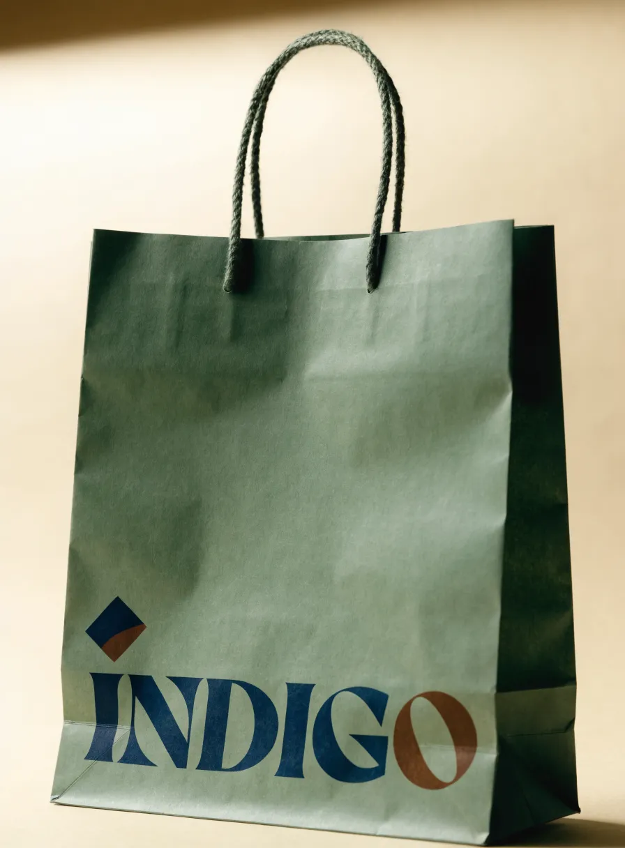 bolsa de papel kraft color verde salvia con asas de cordón torcido y la palabra ÍNDIGO impresa en azul marino con la letra O en cobre, bajo un pequeño rombo azul y cobre, sobre fondo crema con luz cálida