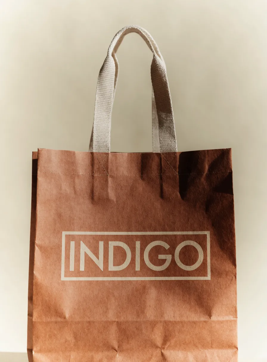 bolsa de papel kraft color terracota con asas planas de algodón y la palabra INDIGO impresa en tinta crema dentro de un marco delgado, sobre fondo crema con luz cálida