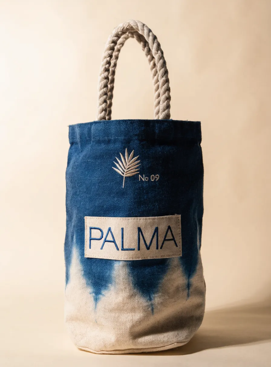 tote cilíndrico de lona teñida a mano en índigo con degradado a crudo, hoja de palma bordada y parche con el nombre de la marca ficticia Palma