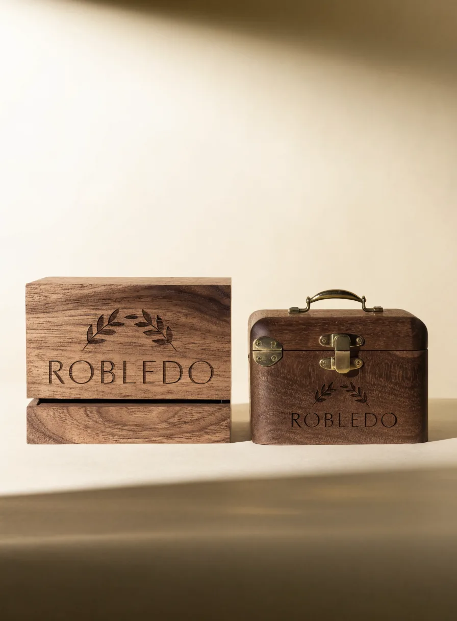 dos cajas de nogal grabadas con la marca Robledo y una corona de laurel, una con tapa corrediza y otra tipo cofre con asa y broche de latón, sobre un fondo color crema