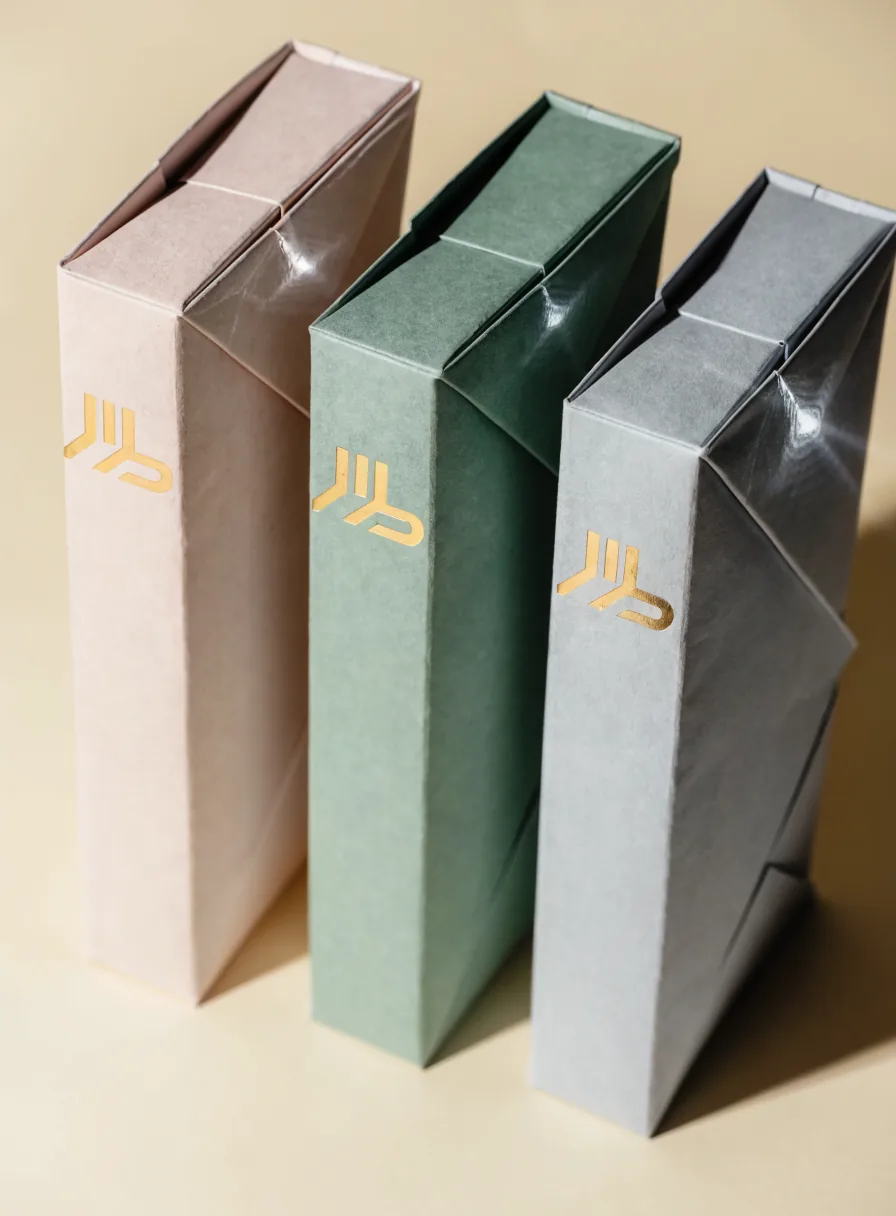 tres estuches plegadizos de pie en blush, verde salvia y gris, cada uno con un emblema geométrico en foil oro, sobre un fondo color crema