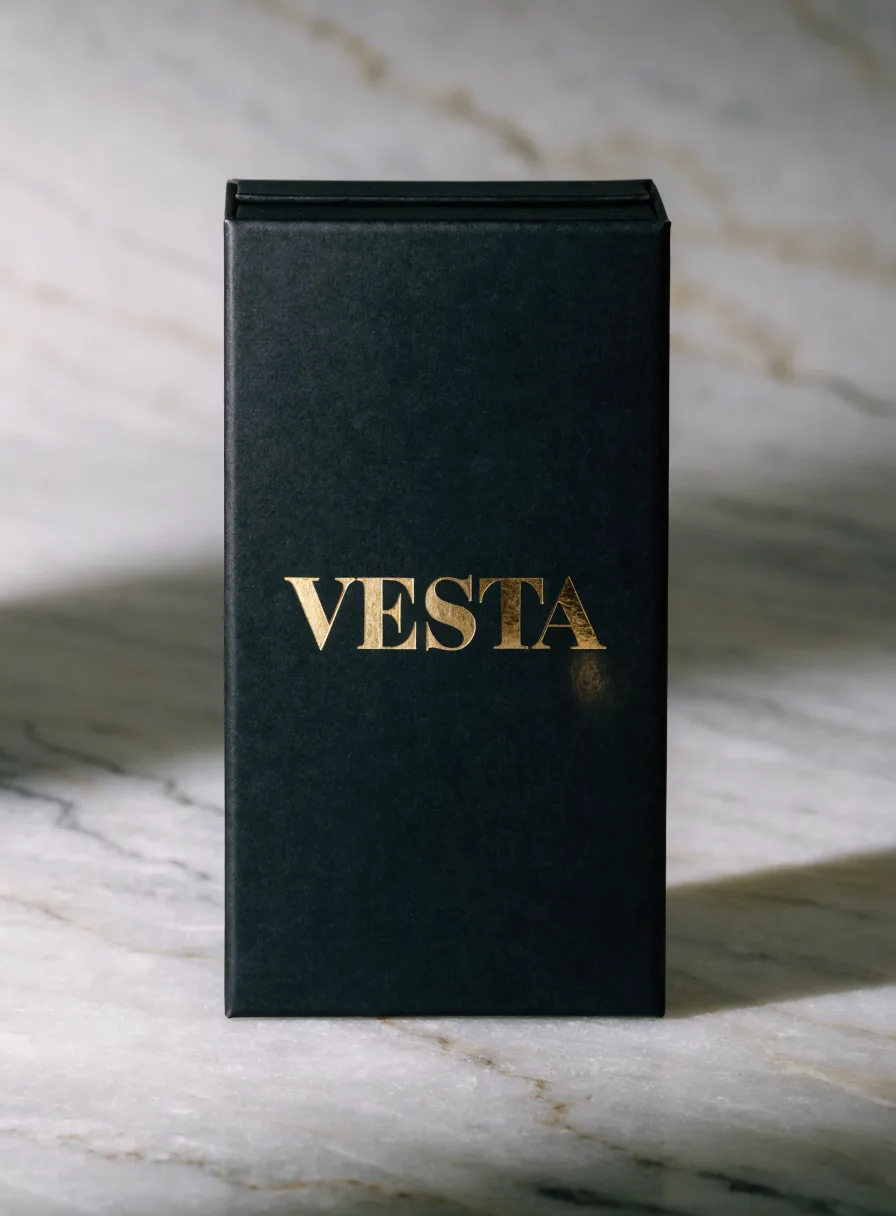 estuche rígido negro mate con la marca ficticia Vesta estampada en foil dorado, sobre una superficie de mármol claro