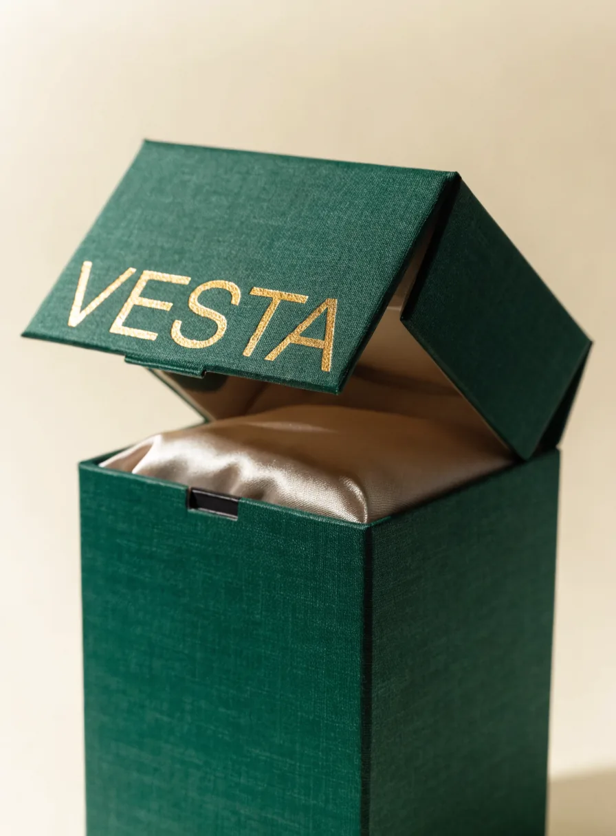 caja rígida imantada forrada en tela verde con el nombre VESTA en foil dorado en la tapa y forro de satín champán adentro