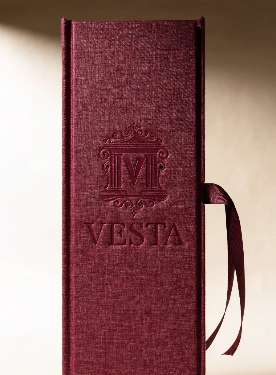 estuche rígido forrado en tela vino con un monograma y el nombre VESTA en bajorrelieve y un listón de tela, de pie bajo luz suave