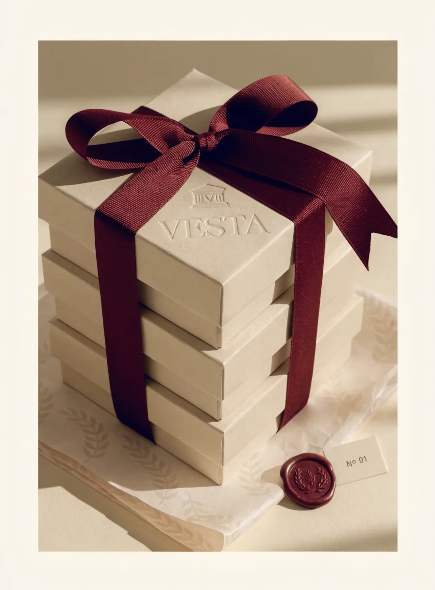 torre de tres cajas rígidas color crema atadas con un listón grosgrain vino en moño, con la marca Vesta en realce y una tarjeta numerada No 01 junto a un sello de lacre