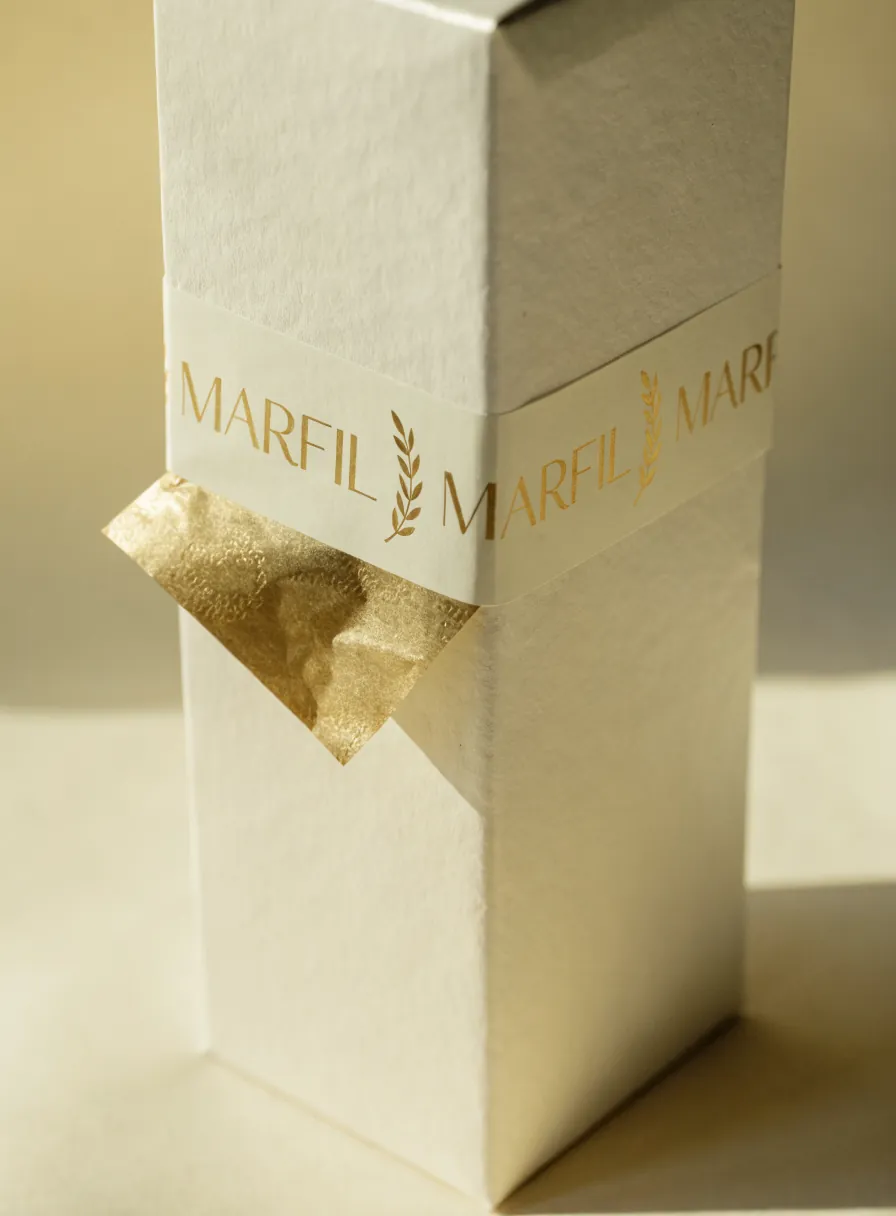 cinta de papel color crema impresa en oro con la marca MARFIL repetida y ramas de laurel, ceñida sobre una caja marfil con papel china dorado asomando
