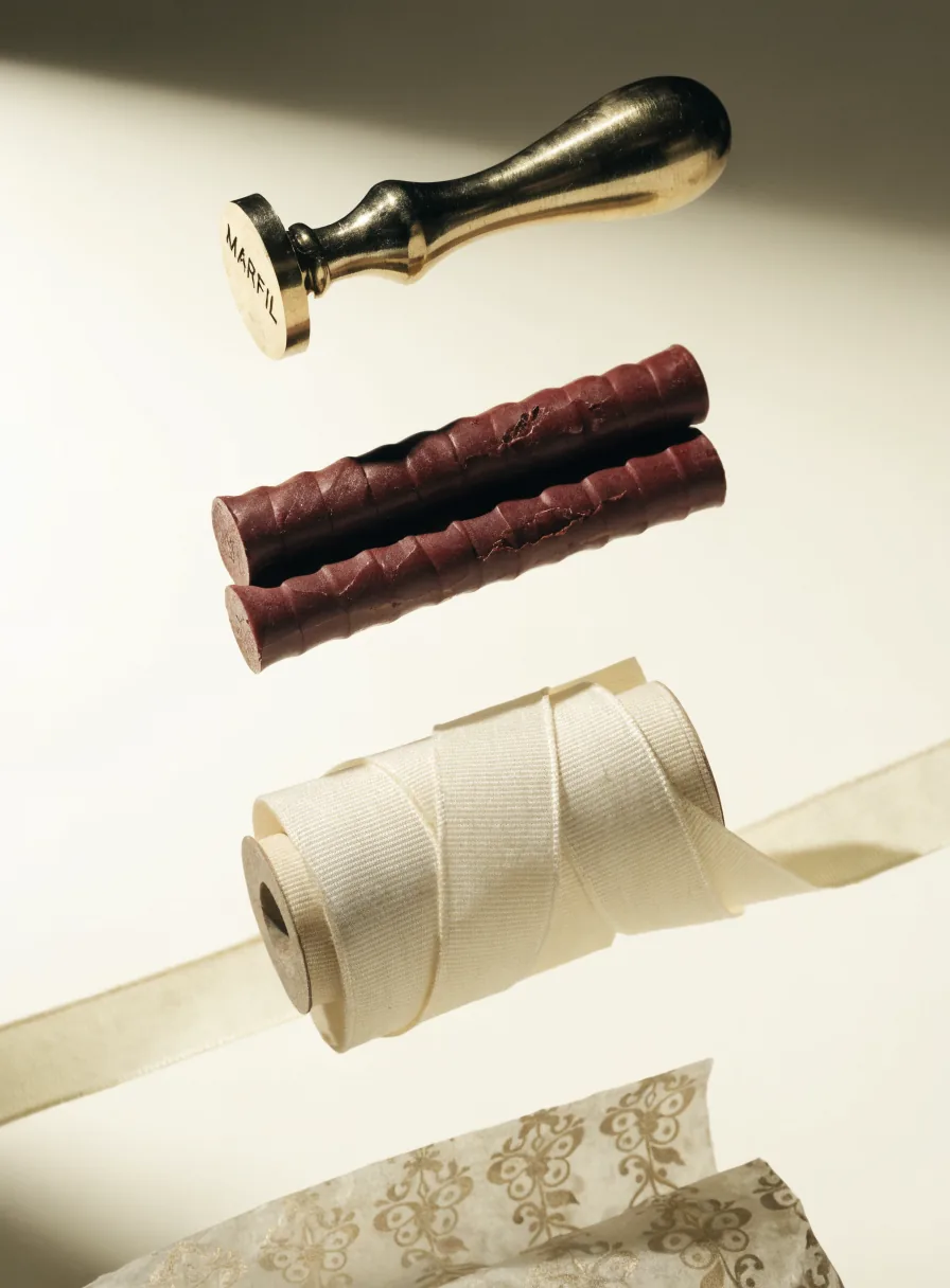 troquel de sellado de latón grabado con la marca MARFIL junto a dos barras de lacre color vino y un carrete de listón marfil sobre papel china dorado