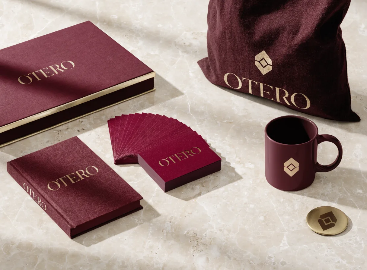 sistema de marca de la firma Otero sobre mármol crema, con caja entelada vino y filo dorado, libreta, tarjetas en abanico, taza, moneda de latón grabada y bolsa de lino, todo con el logotipo en foil dorado