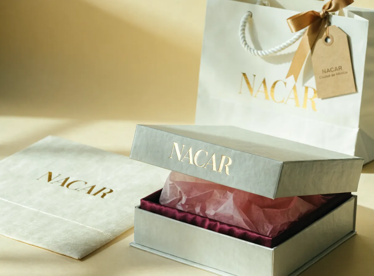 caja rígida color hueso con la marca Nácar en foil oro suave, abierta y con papel de seda e interior de satín magenta, junto a una bolsa de cordón perla con monograma dorado sobre una superficie color hueso