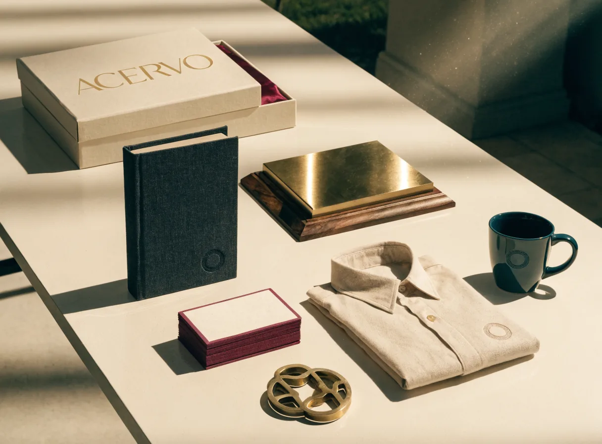 mesa clara de galería con una pieza de cada familia bajo luz de ventana: una caja rígida color hueso con la palabra ACERVO en foil dorado y forro vino, un libro entelado gris carbón de canto, un bloque de tarjetas con el canto pintado en vino, una placa de latón sin grabar sobre base de nogal, una camisa de algodón crema doblada con un emblema bordado al tono, una taza de cerámica azul petróleo y un emblema circular calado en latón