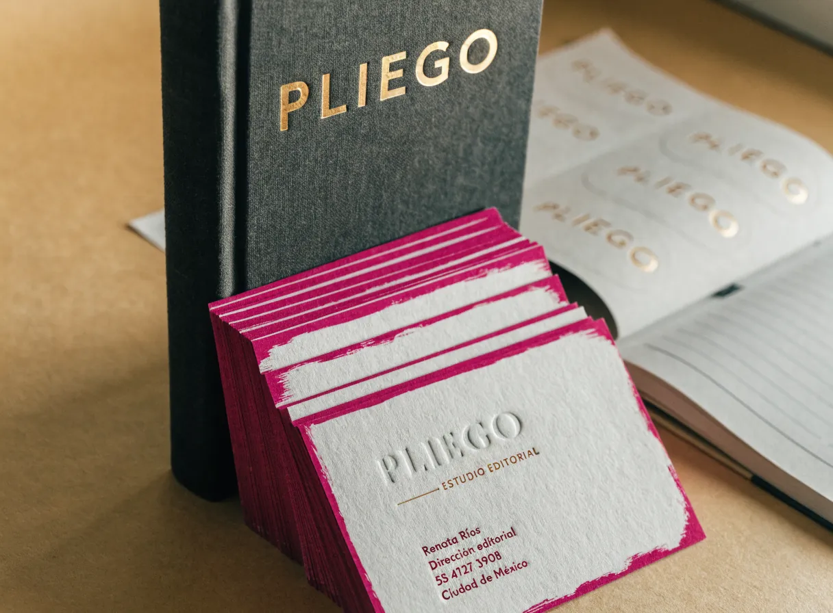 un libro entelado en gris carbón con la palabra PLIEGO en foil dorado, un abanico de tarjetas de algodón con el canto pintado de magenta y el nombre del estudio ficticio PLIEGO en letterpress, una hoja de etiquetas troqueladas y una agenda abierta sobre papel kraft