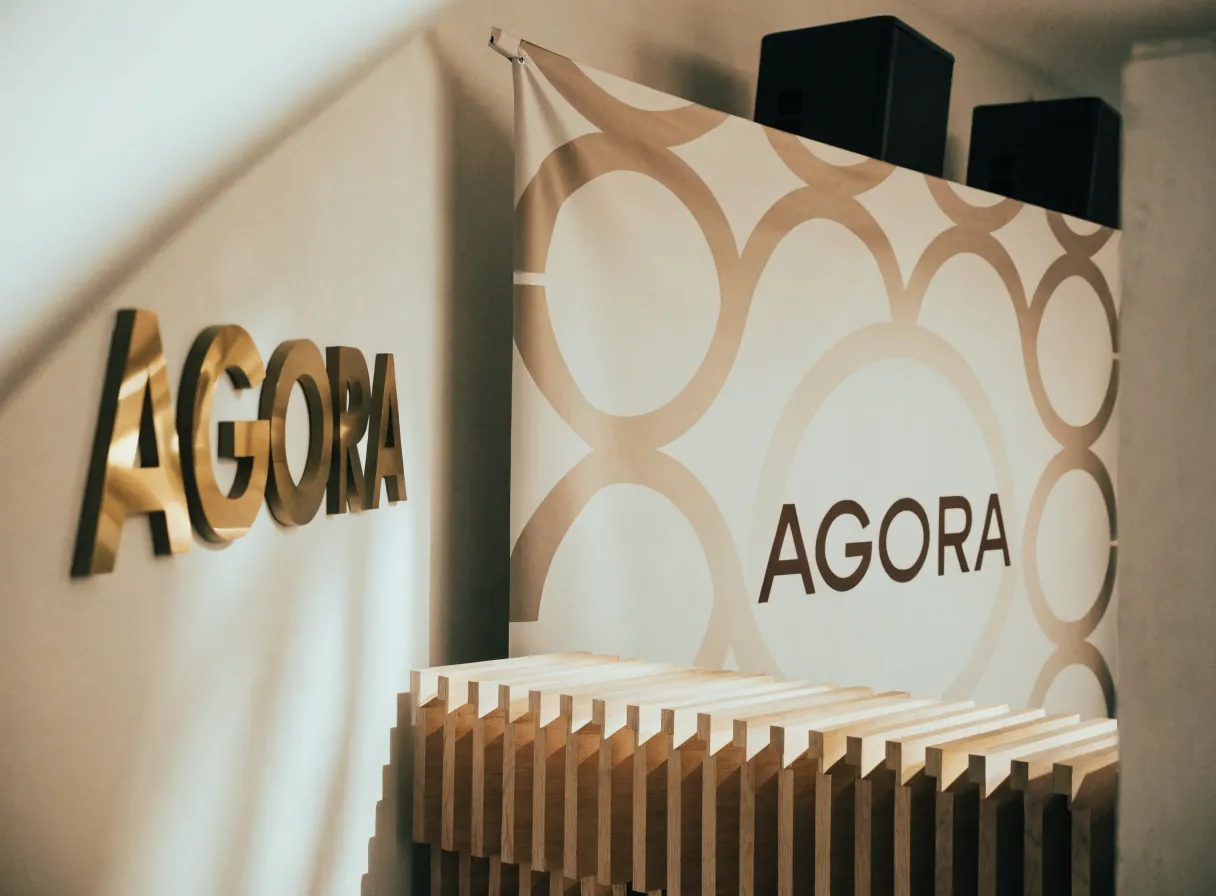montaje de activación de marca con letras corpóreas de latón AGORA sobre el muro, un backdrop impreso con patrón de círculos entrelazados y una banca baja de listones de madera al frente, bajo luz cálida de salón