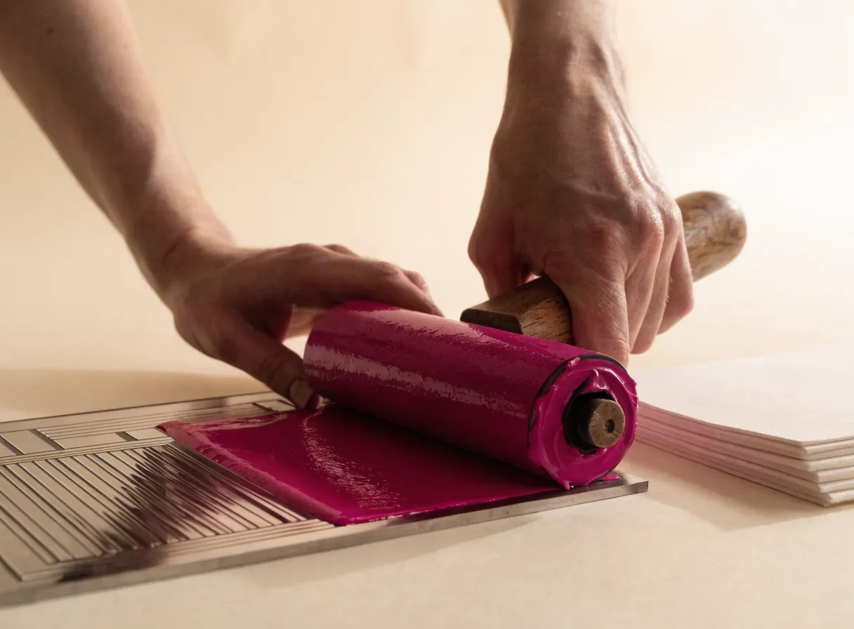 manos entintando una plancha de patrón geométrico con un brayer en tinta magenta