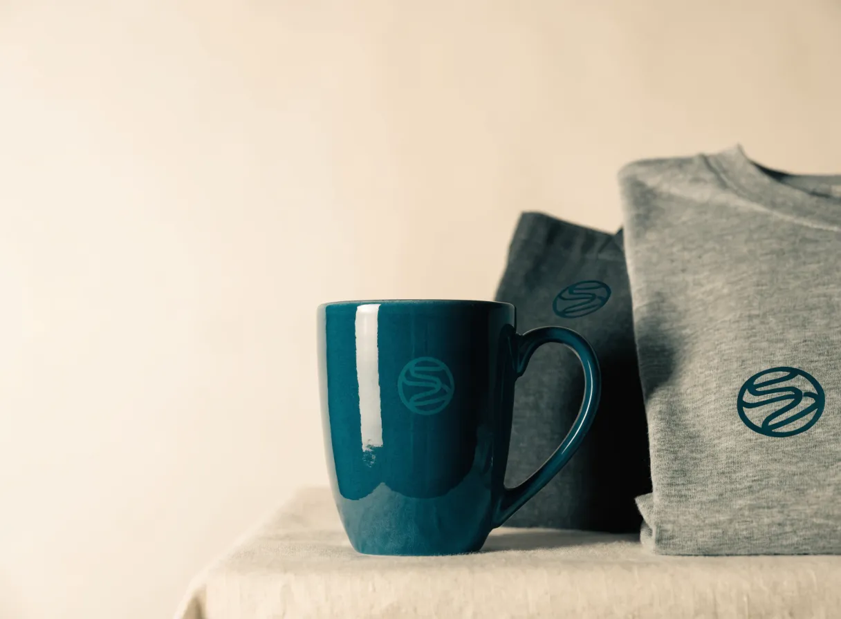 taza de cerámica teal y playera gris con la marca de la empresa, en juego sobre lino claro