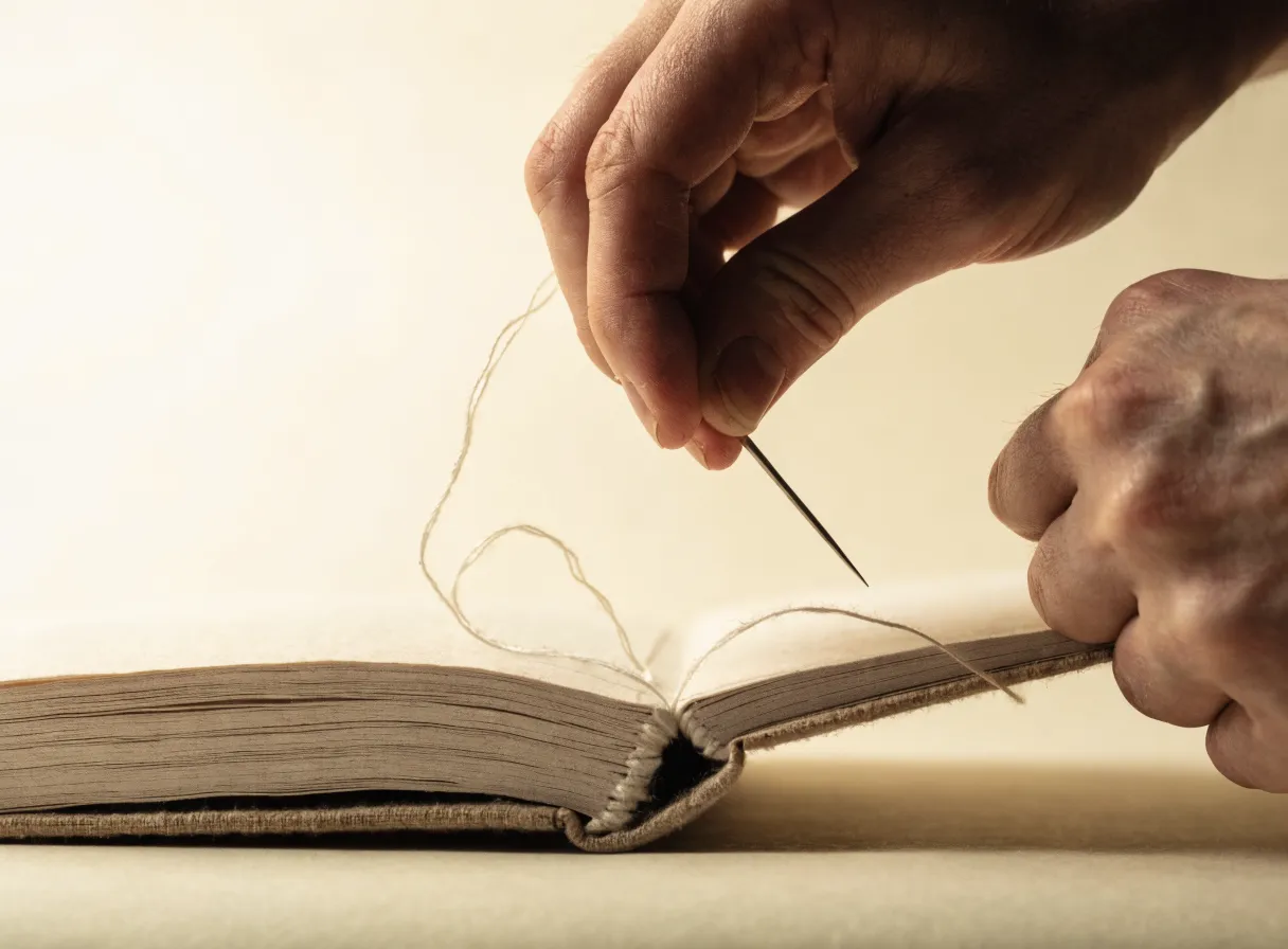 dos manos cosen a mano el lomo de un libro abierto con aguja e hilo de lino sobre una mesa de trabajo clara