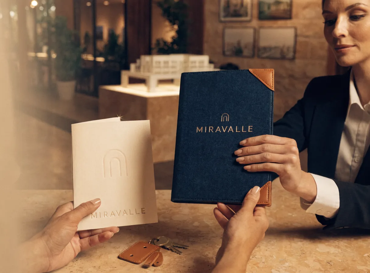 asesora inmobiliaria entrega la carpeta de cierre de piel azul marino de la marca Miravalle a las manos del comprador en la sala de ventas, con un llavero de piel y las llaves sobre la mesa de travertino