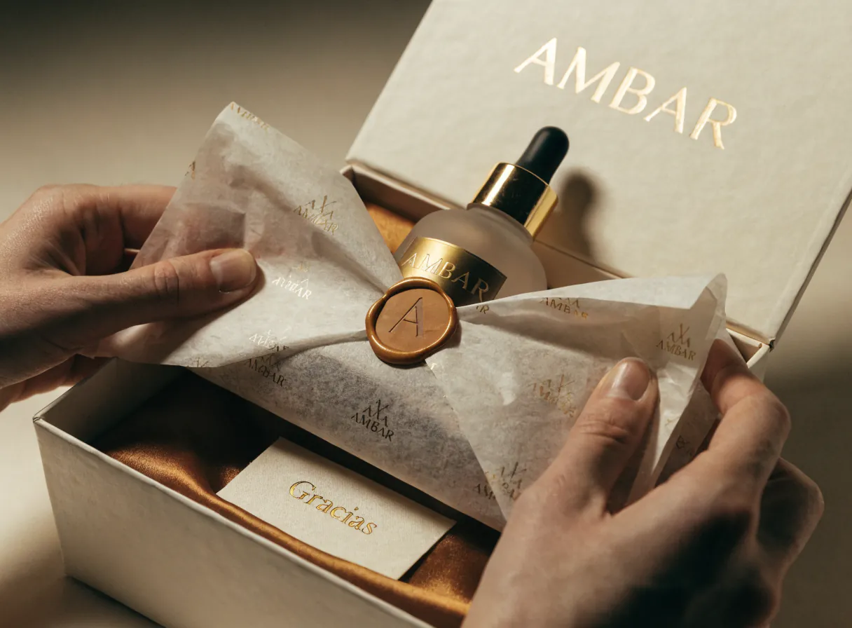 dos manos abren el papel de seda con el monograma de la marca ficticia AMBAR sobre un sello de cera, y aparece un frasco gotero de vidrio esmerilado con cuello dorado dentro de una caja rígida color hueso con interior de satín y una tarjeta que dice Gracias