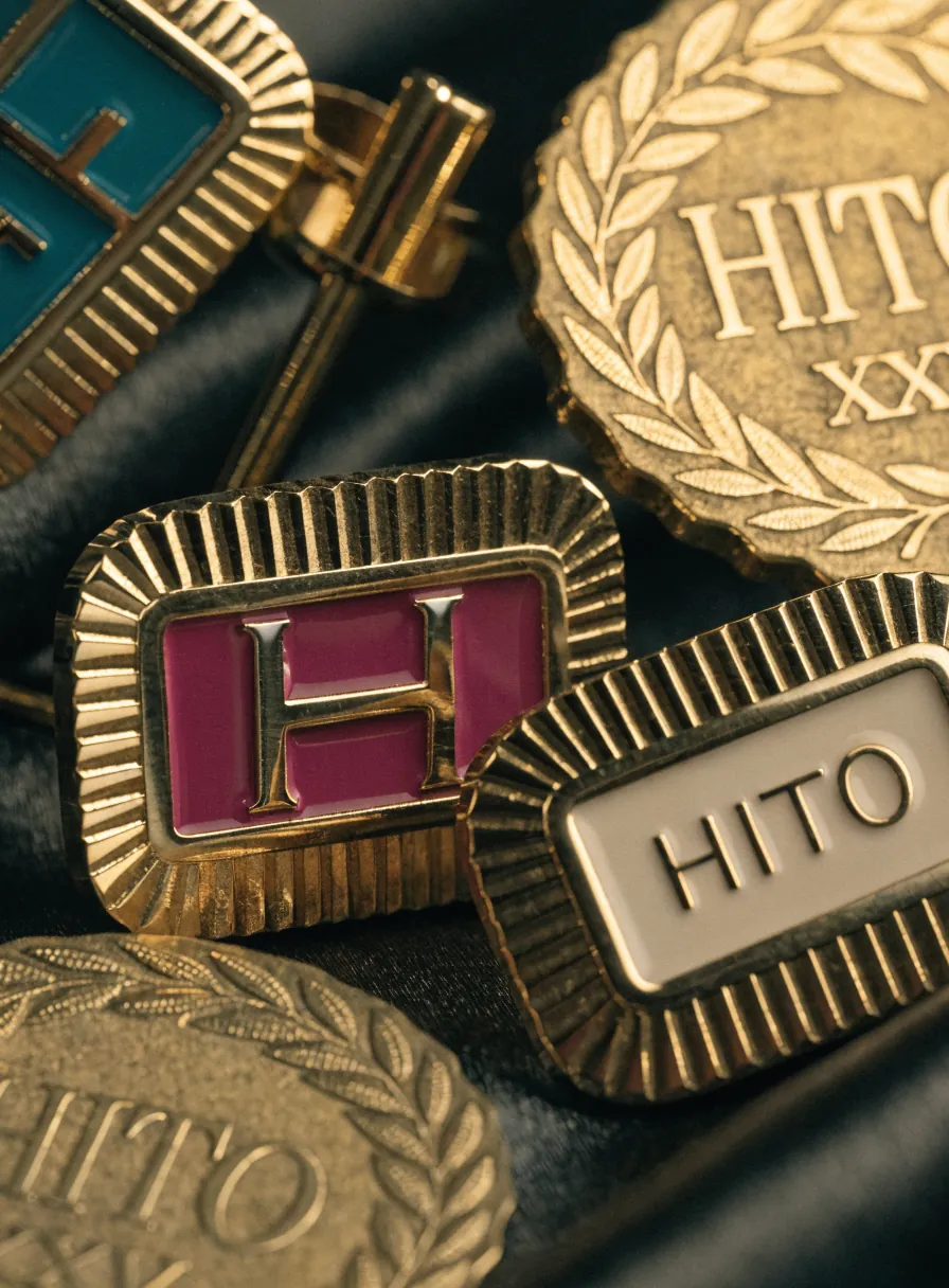 pines de solapa en latón dorado con esmalte, uno con un monograma H sobre esmalte magenta y otro con la palabra HITO sobre esmalte marfil, junto a medallas acuñadas con laurel sobre tela oscura