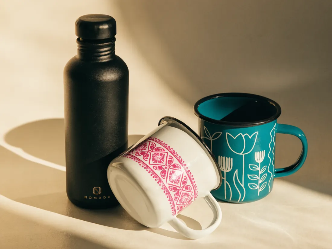 botella térmica negra con la marca NOMADA junto a dos tarros de peltre, uno blanco con patrón geométrico rosa y otro teal con flores, ambos con diseño envolvente a todo color