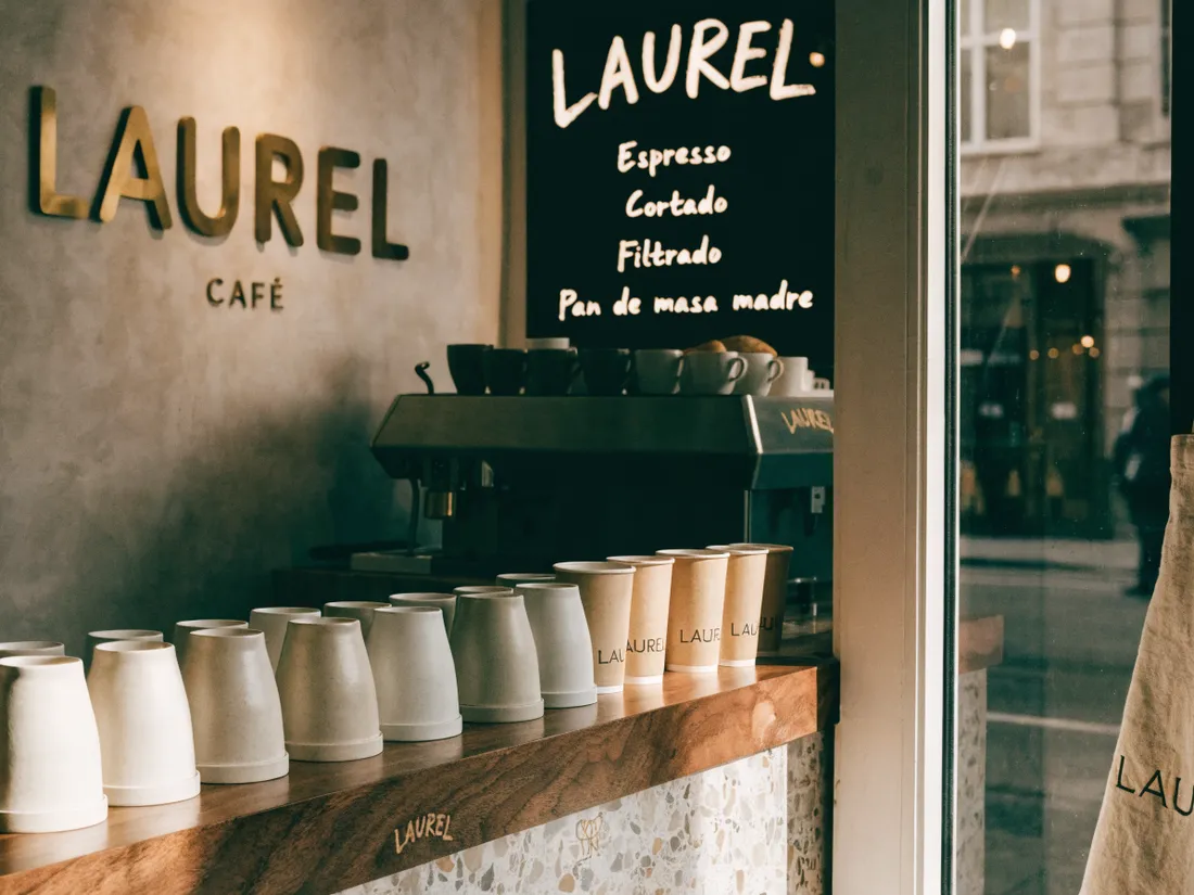 interior del café de muestra LAUREL con letras corpóreas doradas de latón montadas en el muro de cemento, pizarra de menú y tazas de la marca alineadas en la barra de madera y terrazo