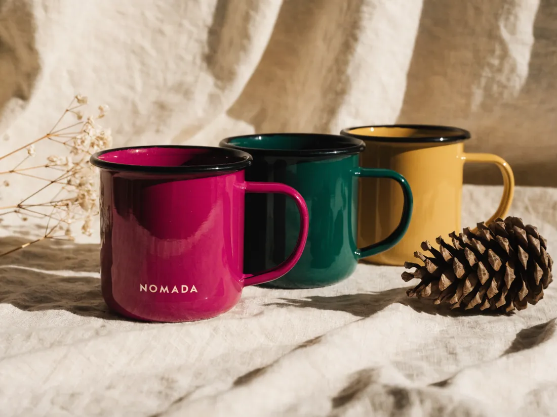 tres tazas de peltre esmaltado en magenta, verde bosque y mostaza con la marca Nomada, junto a una piña de pino sobre lino natural