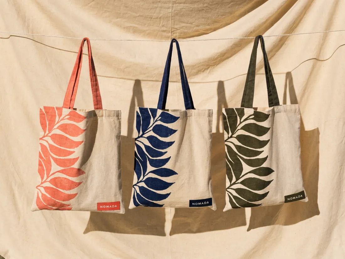 tres totes de lona natural con un motivo de hoja serigrafiado en coral, azul marino y verde olivo, y la marca ficticia Nómada en una etiqueta
