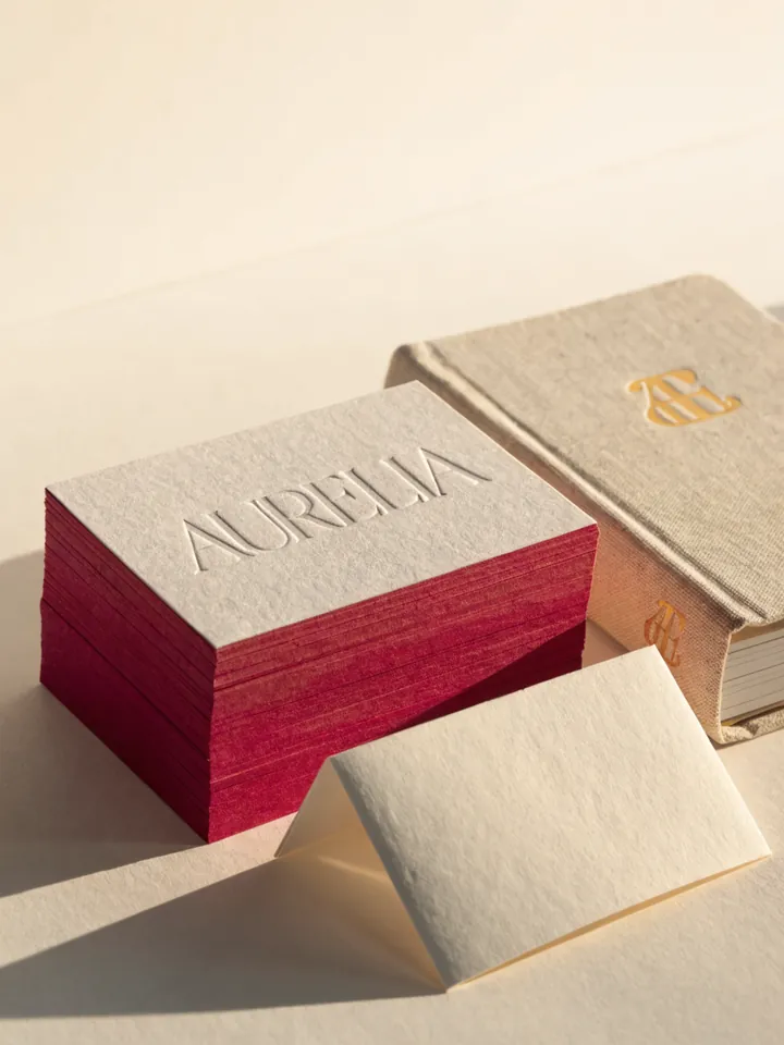 bloque de tarjetas de algodón con canto pintado en rojo y el nombre en relieve al seco, junto a un sobre y una libreta de lino con monograma dorado