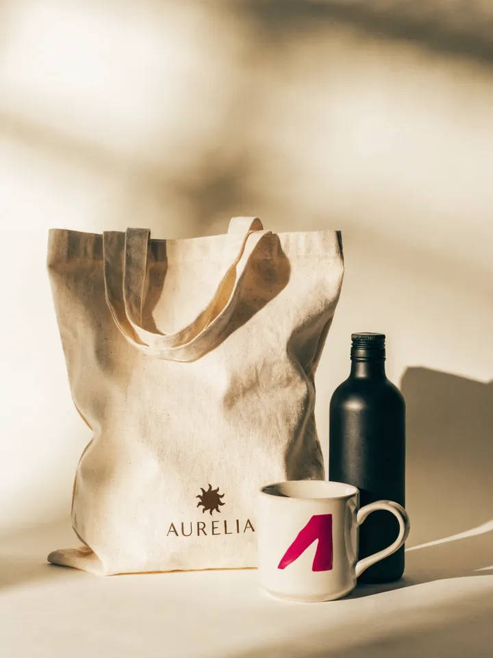 kit de merch en fondo crema con un tote de algodón, una taza blanca y un termo negro, todos con la marca AURELIA impresa