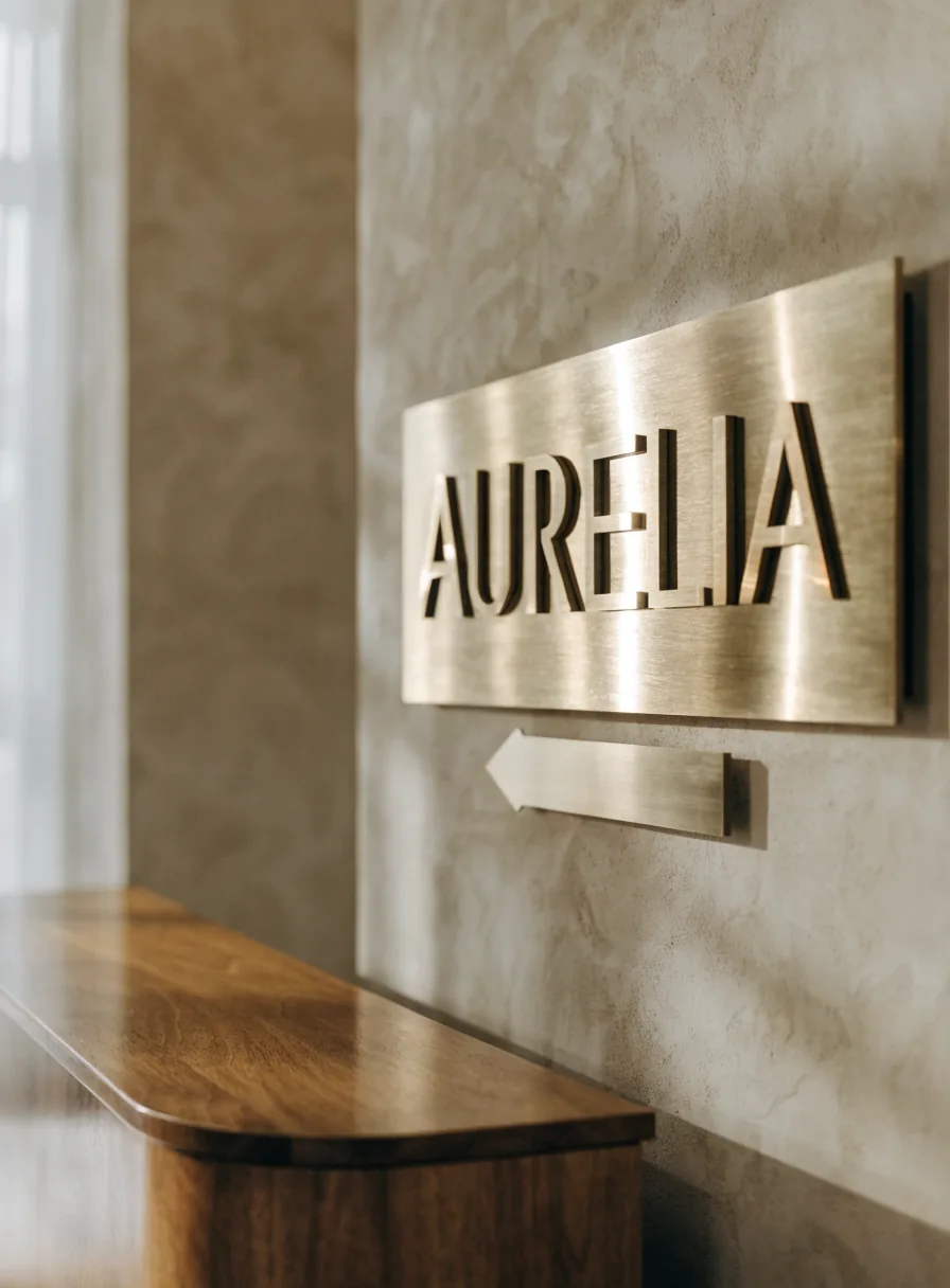Placa de latón cepillado con letras caladas AURELIA montada sobre muro de yeso cálido, con su plaquita direccional