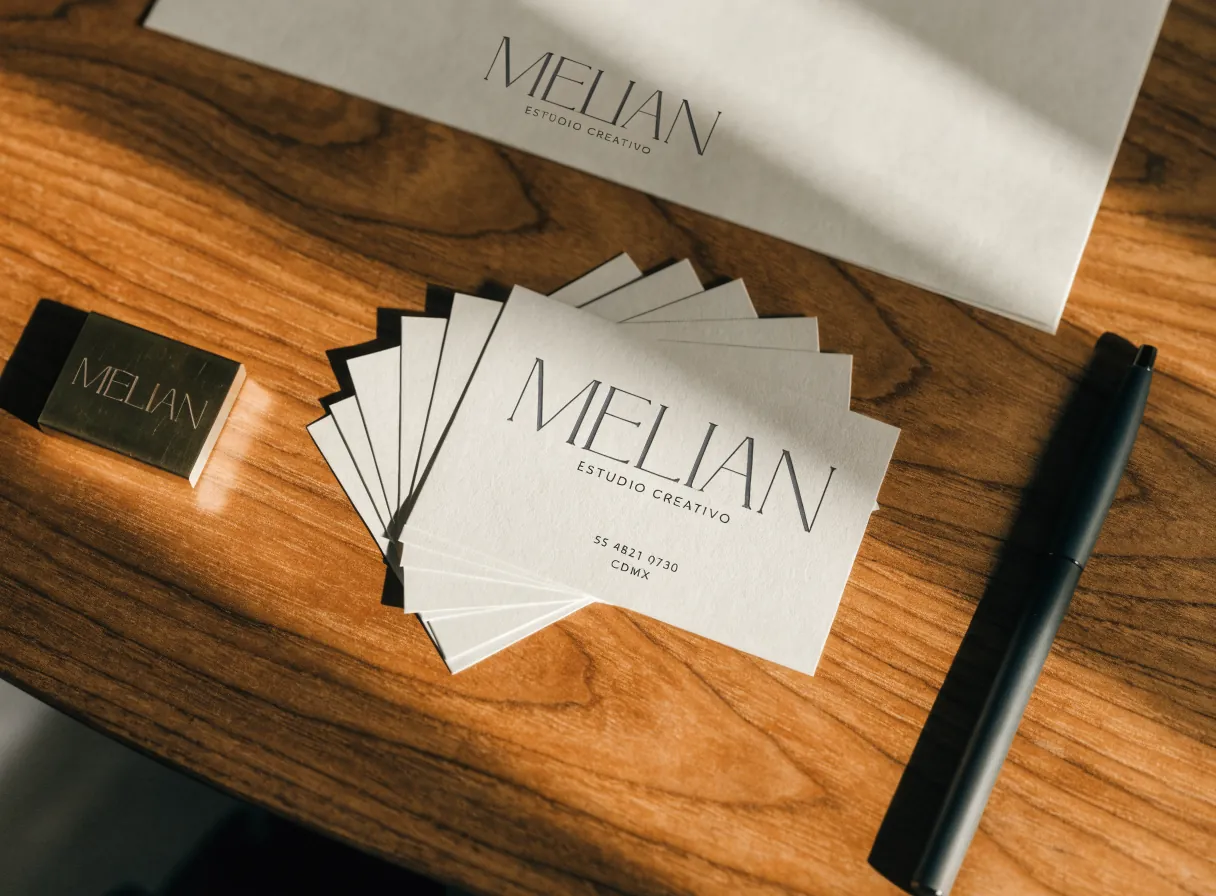 un juego de tarjetas de presentación en abanico junto a una hoja membretada y un porta-tarjetas de latón grabado con la marca Melián sobre un escritorio de nogal, con luz de ventana por la mañana
