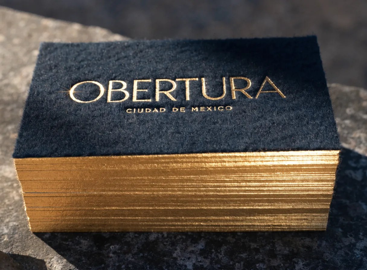 bloque de tarjetas de presentación azul noche de algodón con la marca Obertura en foil dorado a fuego y el canto pintado en oro, sobre piedra oscura