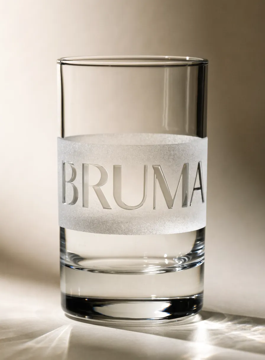 Vaso alto de cristal grueso en vertical, con una banda arenada a media altura y el nombre BRUMA reservado en pulido, iluminado por luz cálida lateral.