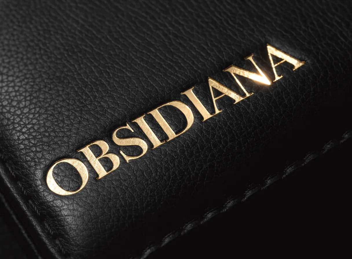 Esquina de una carpeta de piel negra granulada con un nombre estampado en foil dorado, iluminada de lado para que el relieve del cuño levante su propia sombra.
