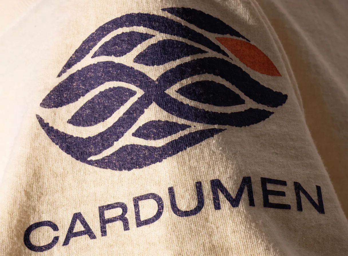 acercamiento al hombro de una playera de algodón crudo con un emblema tejido azul marino y un acento naranja serigrafiado a mano y la palabra CARDUMEN debajo