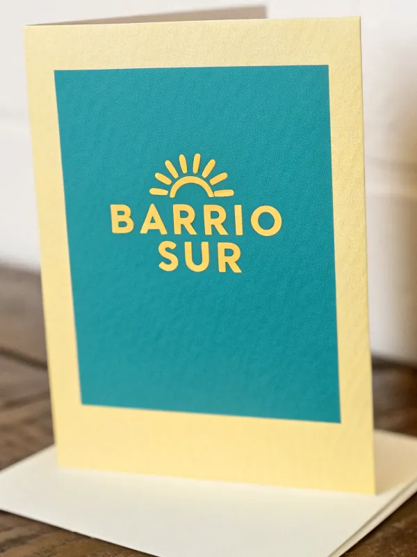 tarjeta doblada de papel crema de pie sobre su sobre blanco, con un panel turquesa serigrafiado y encima un sol y la palabra BARRIO SUR en amarillo