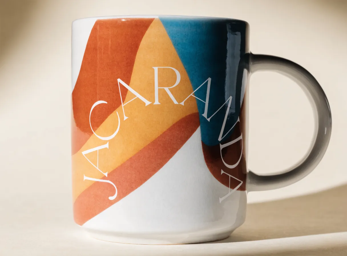 Taza de cerámica blanca esmaltada con bandas curvas en terracota, ámbar y azul petróleo y una palabra en blanco cruzando en diagonal.