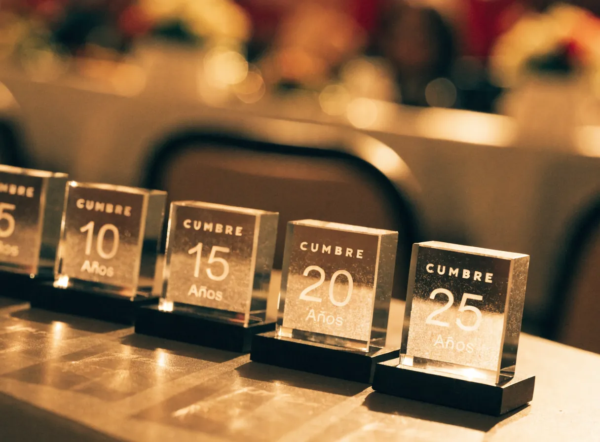 cinco reconocimientos de cristal alineados sobre la mesa larga de un salón, grabados CUMBRE con 5, 10, 15, 20 y 25 años, bajo la luz cálida del banquete y con los centros de mesa desenfocados al fondo