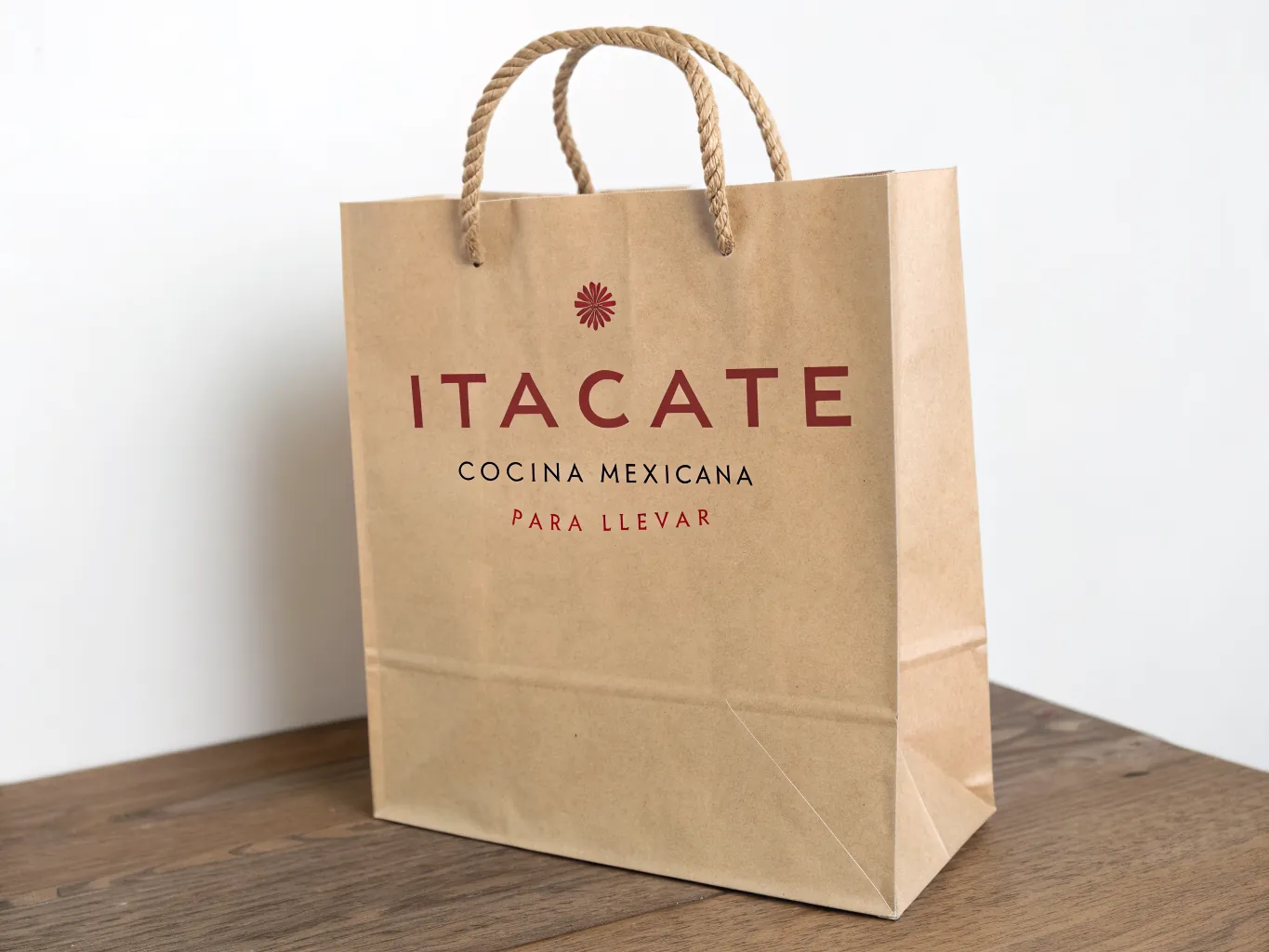 bolsa de papel kraft con asas de cordón grueso y la marca ITACATE impresa en rojo, con la leyenda cocina mexicana para llevar en negro, sobre una mesa de madera
