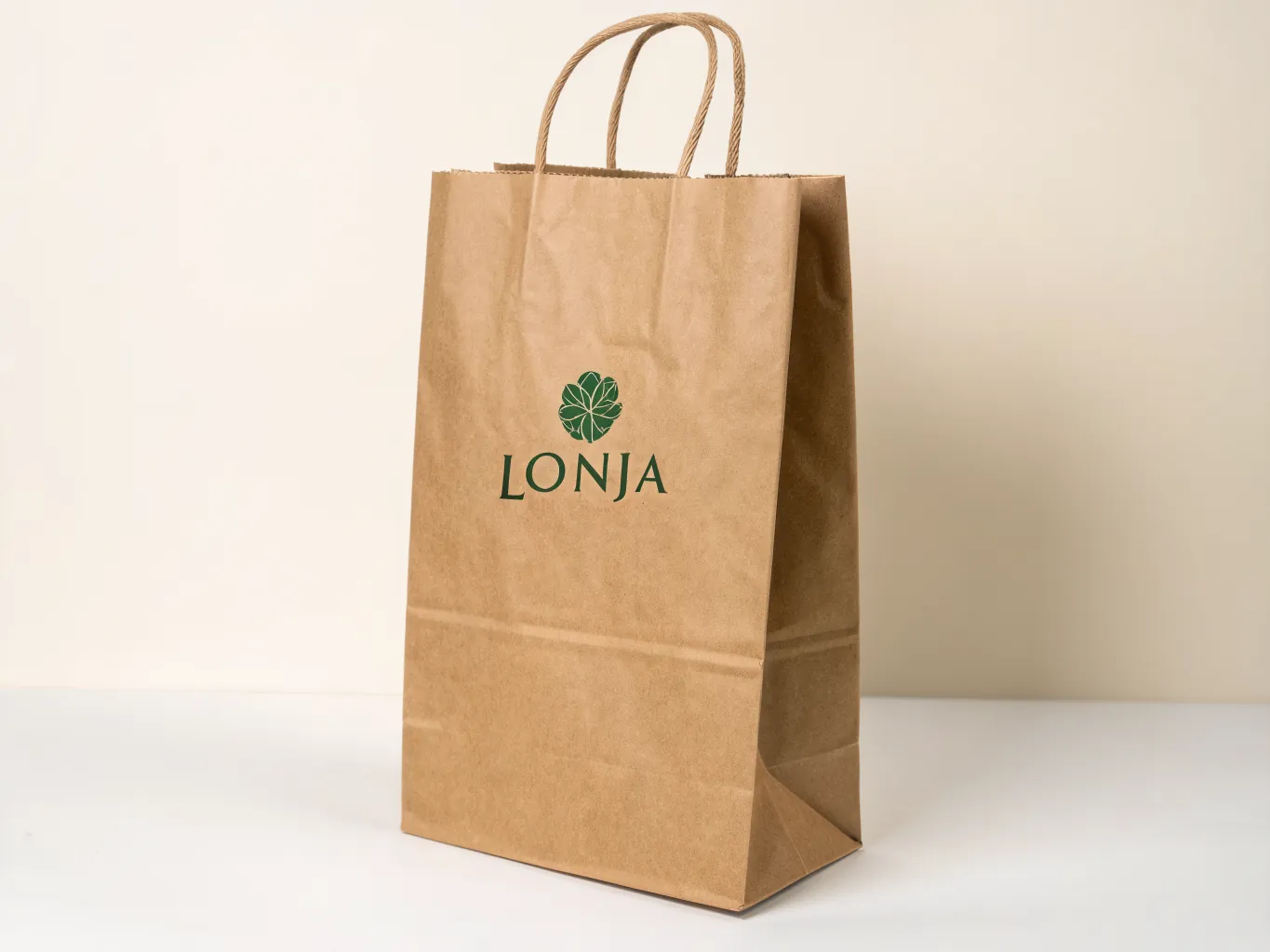 bolsa de papel kraft con asas de papel torcido y la marca LONJA impresa en verde a una tinta bajo una flor de hojas, sobre fondo crema
