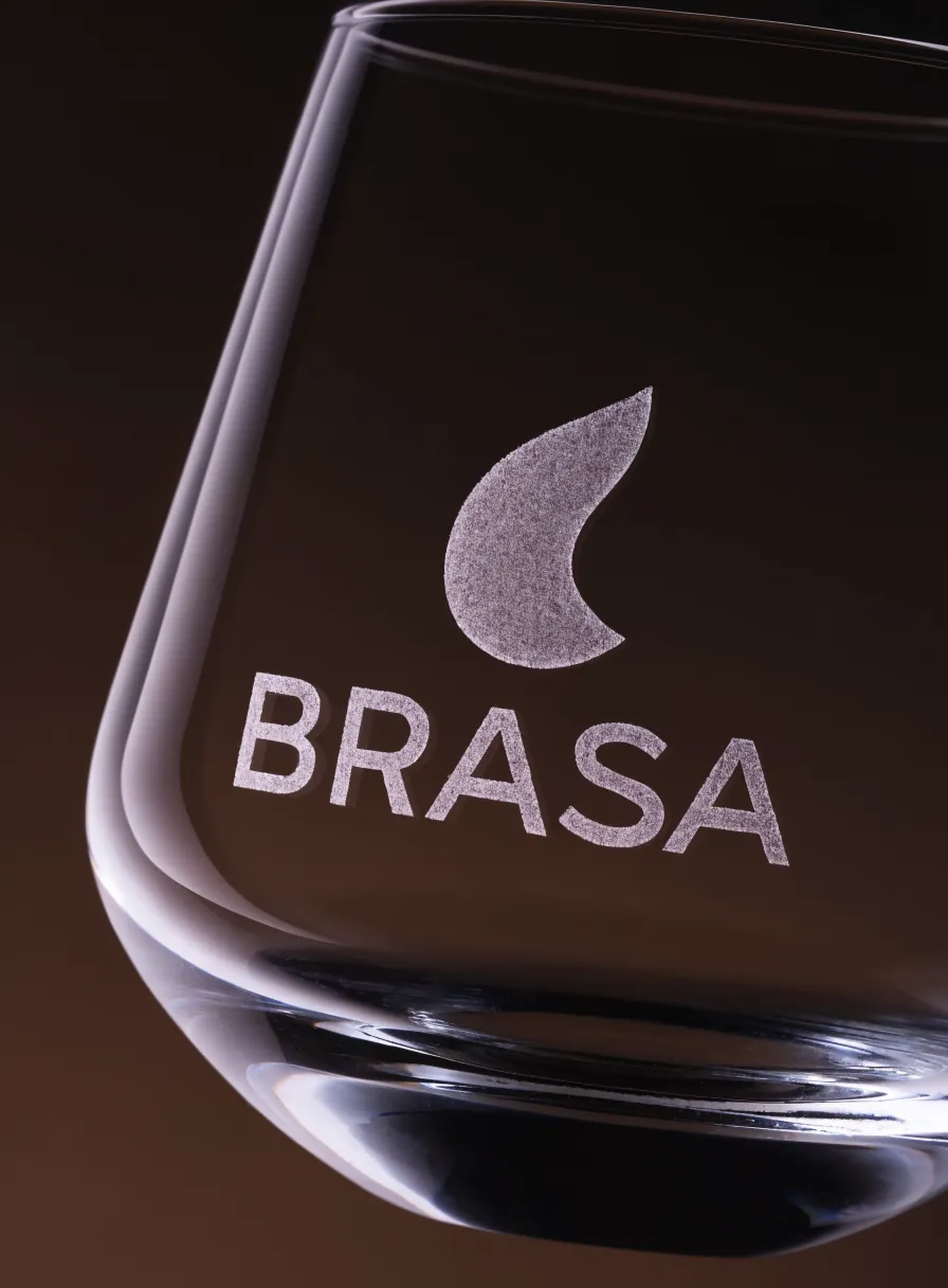 copa de cristal sin tallo con la marca BRASA y una gota arenada, sobre un fondo cálido oscuro
