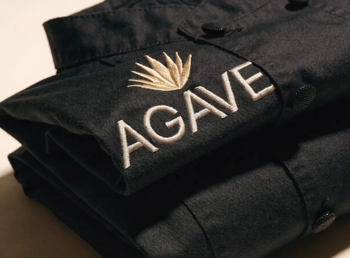 pila de filipinas de chef en algodón negro dobladas una sobre otra, con la de encima mostrando un agave bordado en hilo dorado y la palabra AGAVE bordada en crema debajo
