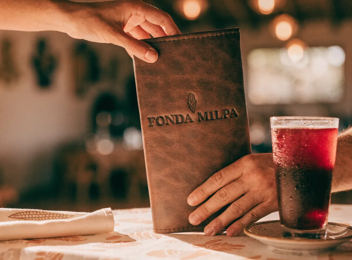 unas manos entregan un portamenú de piel café con el nombre FONDA MILPA y una mazorca hundidos en seco, sobre una mesa con mantel bordado y un vaso de agua de jamaica, con el bar del fondo desenfocado