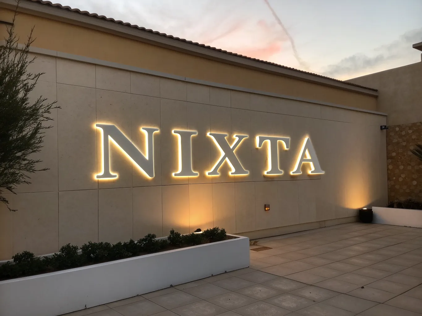 Letras corpóreas blancas de gran tamaño con el nombre NIXTA montadas sobre una fachada de piedra clara, retroiluminadas al atardecer con un halo tibio derramado sobre el muro.