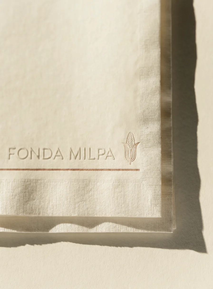servilleta de papel color crema con la marca Fonda Milpa y una espiga de maíz en relieve seco, sobre una fina línea impresa en terracota