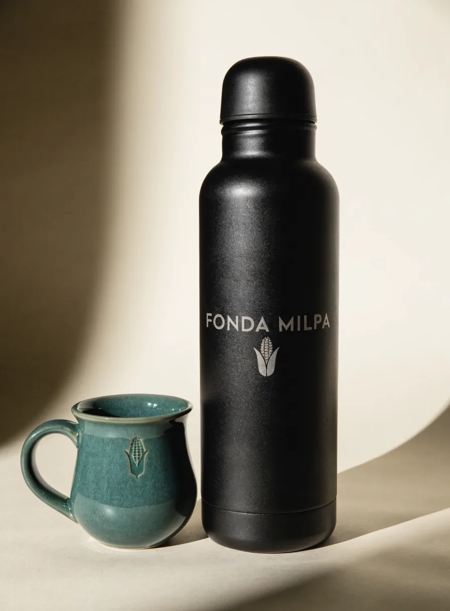 termo de acero mate negro con la marca Fonda Milpa y una mazorca grabadas a láser, junto a una taza de cerámica verde a juego